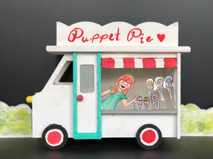 A small birdhouse painted to look like an ice cream truck. Paper puppets of stacey and ice cream puppets are inside the window. The truck is white with a red and white awning and a teal door.