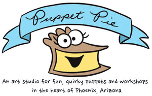 An Art Studio for for fun quirky puppets and workshops in the heart of Phoenix, Arizona. Photo is of a blue banner with the words Puppet Pie in the banner. It is magically floating over a piece of custard pie that has eyeballs and is smiling